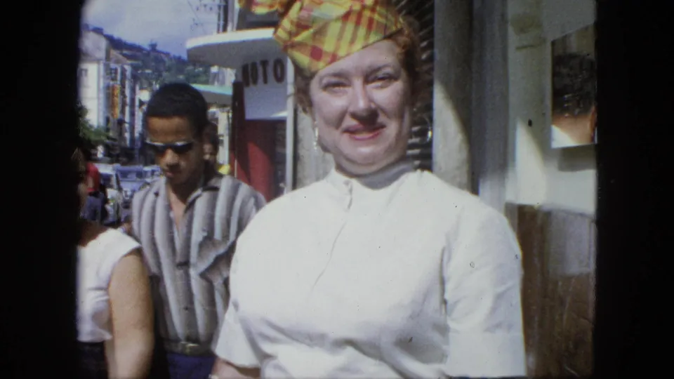 A close handheld frame shows the smiling woman in a white dress near a storefront edge, with pedestrians and cars behind her.
