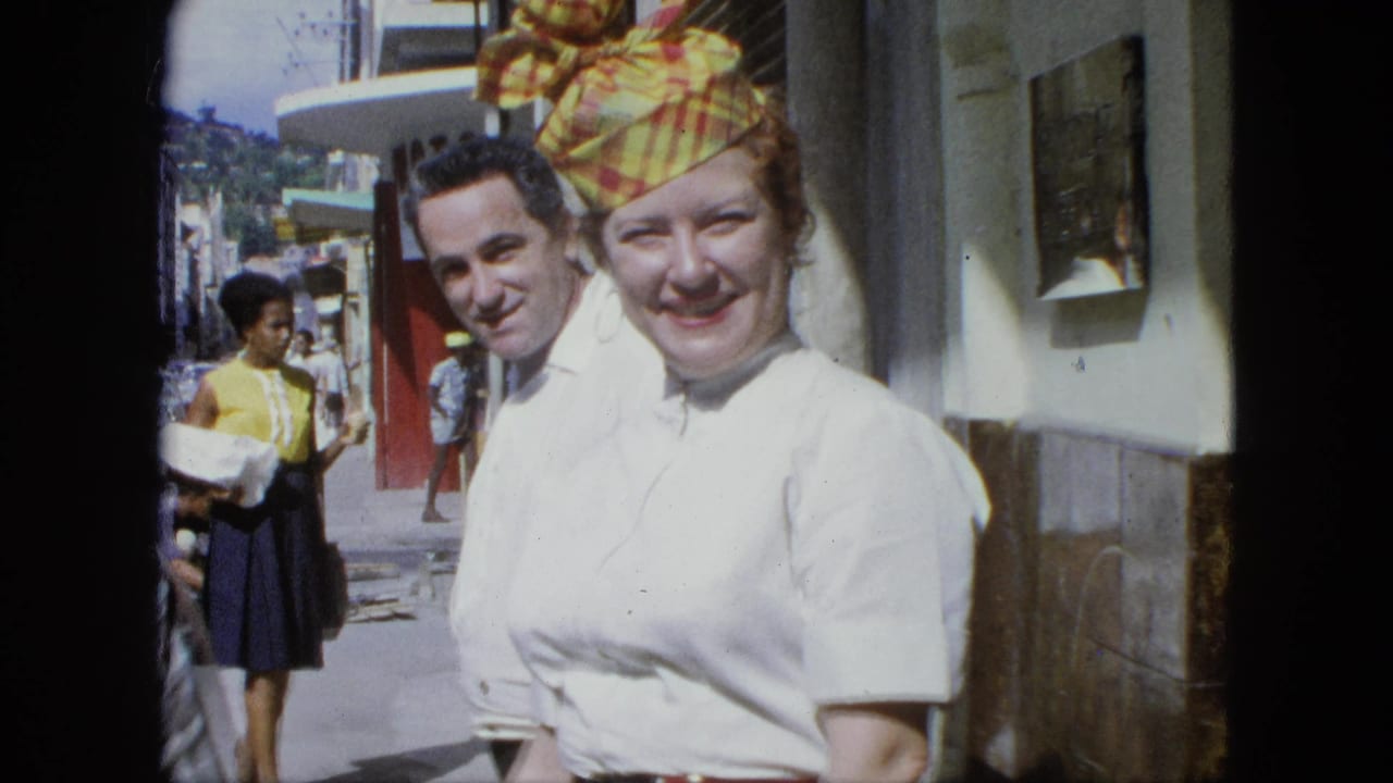1964 U.S. Virgin Islands frame of a smiling woman in a white blouse and colorful headscarf, with a man and pedestrians behind her.