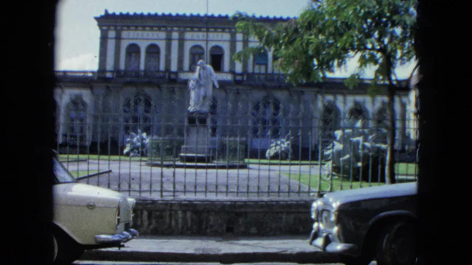 1964 U.S. Virgin Islands home-movie view of a formal building behind a wrought-iron fence, with a statue, trees, and parked cars.