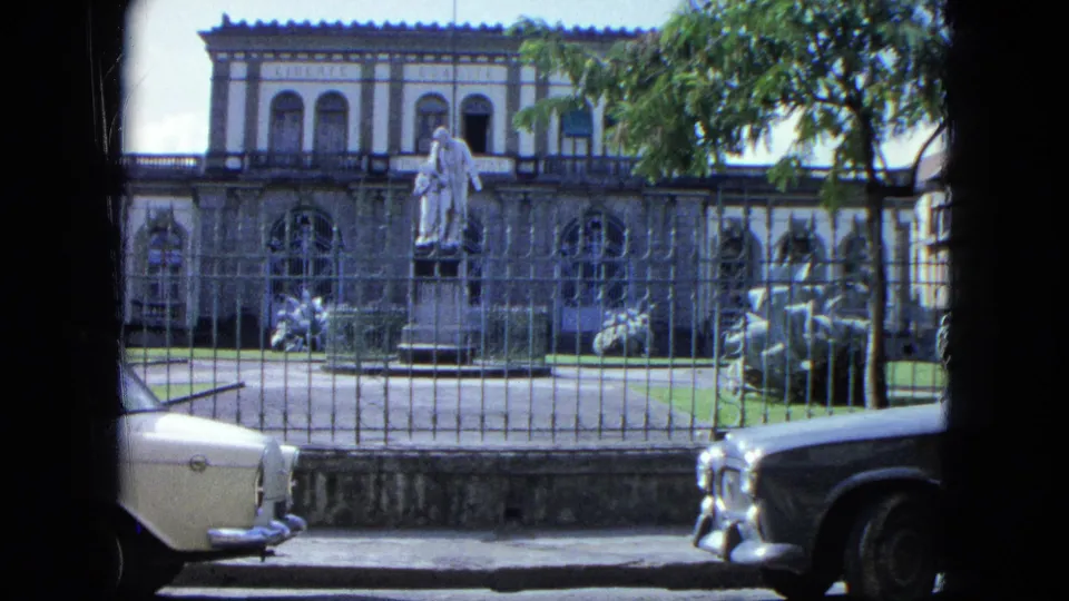 1964 U.S. Virgin Islands frame showing a public-looking building with a statue behind fencing, partially shaded by trees and bordered by parked cars.
