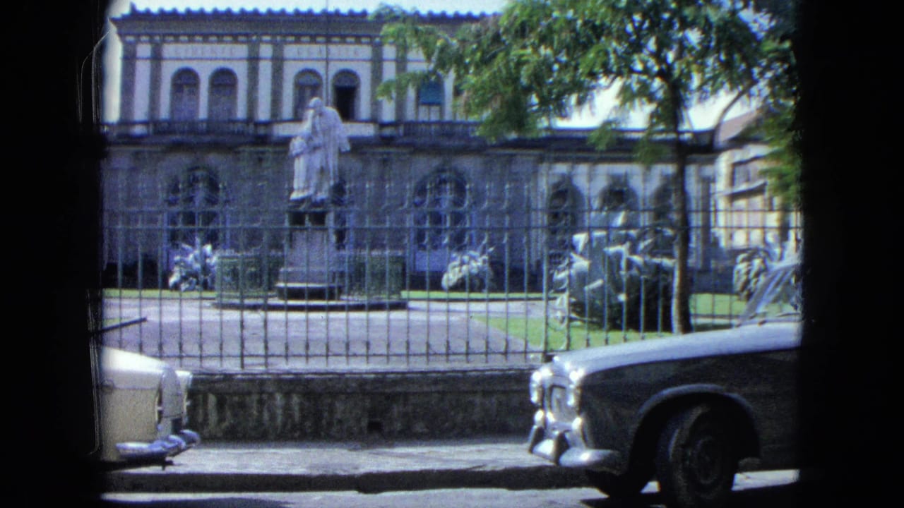 Colonial-style building with statue behind an iron fence, seen past parked classic cars.