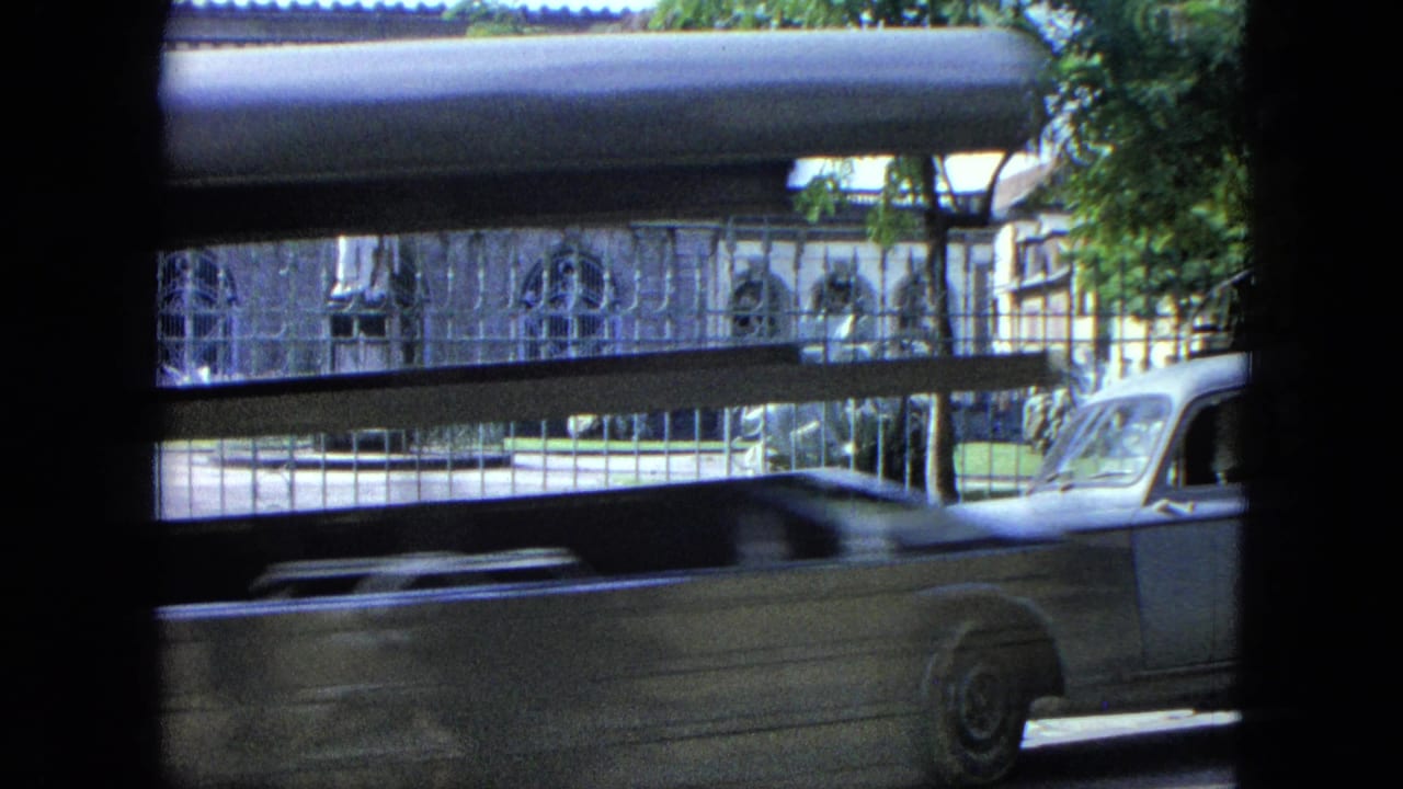 A 1964 USA Virgin Islands street scene filmed through a vehicle window, with a fenced building, statue, and light classic pickup passing by.