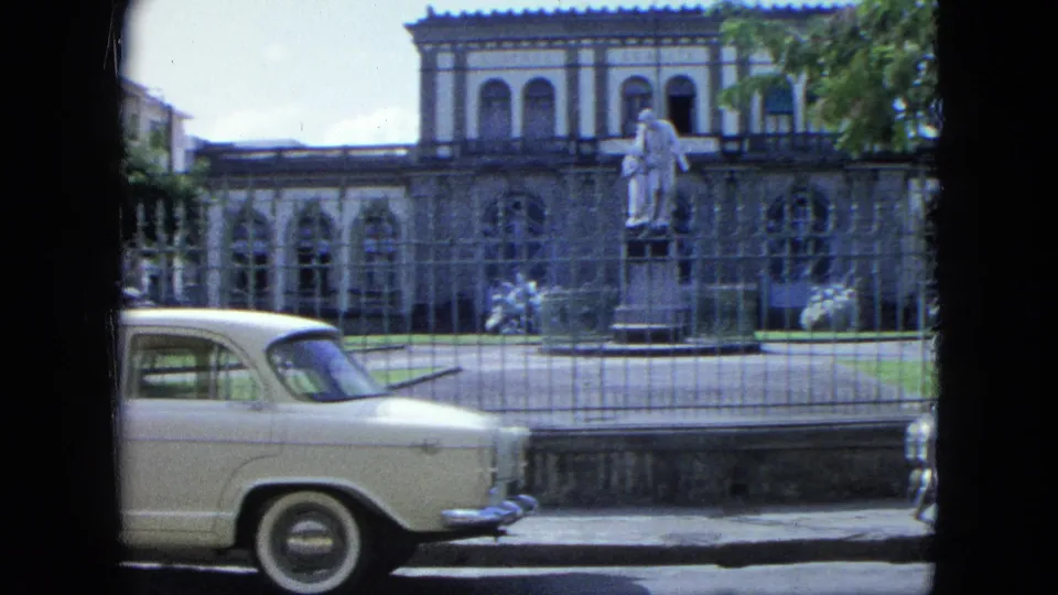 A 1964 USA Virgin Islands street frame shows a white classic sedan passing a fenced colonial-style building with a central statue.
