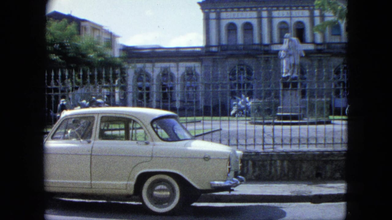 White classic car parked beside an iron fence in front of an arched building and statue.