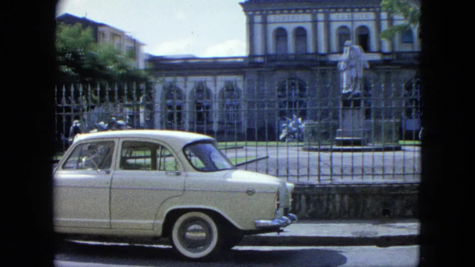 1964 home-movie frame of a white classic car along a street, with an iron fence, statue, and arched building behind it.