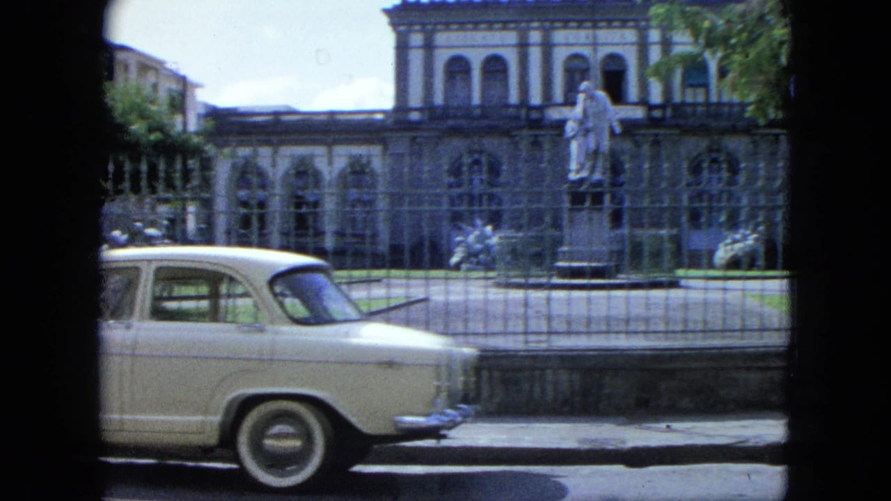 White classic car framed beside a fenced building with arched windows and a statue.