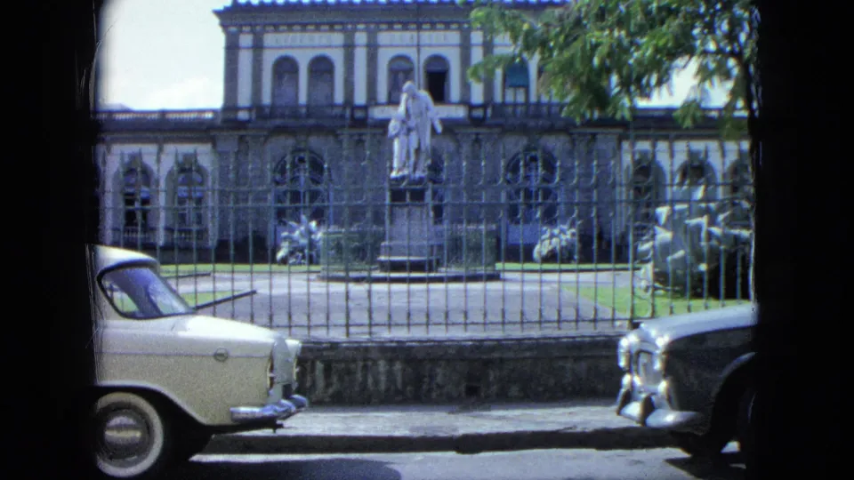 Visitor-shot street frame with a partial white car, another car entering, iron fence, statue, and arched building facade.