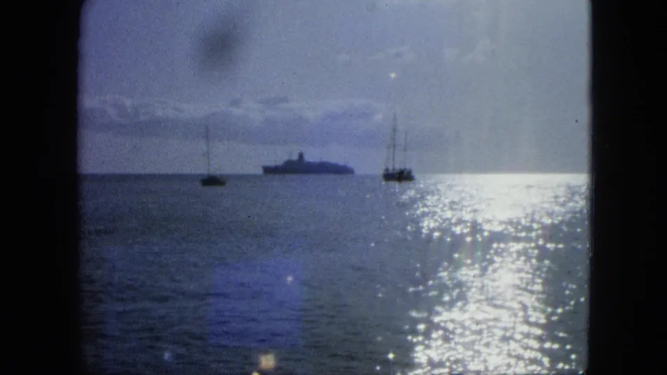 Coastal 8mm frame showing sparkling ocean water, two small sailboats, and a larger distant vessel under clouds.