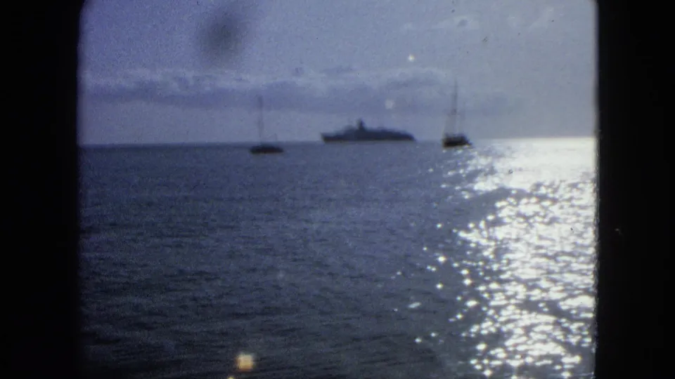 Soft 8mm frame from the 1964 U.S. Virgin Islands clip showing offshore boats and a larger vessel under cloudy sky.