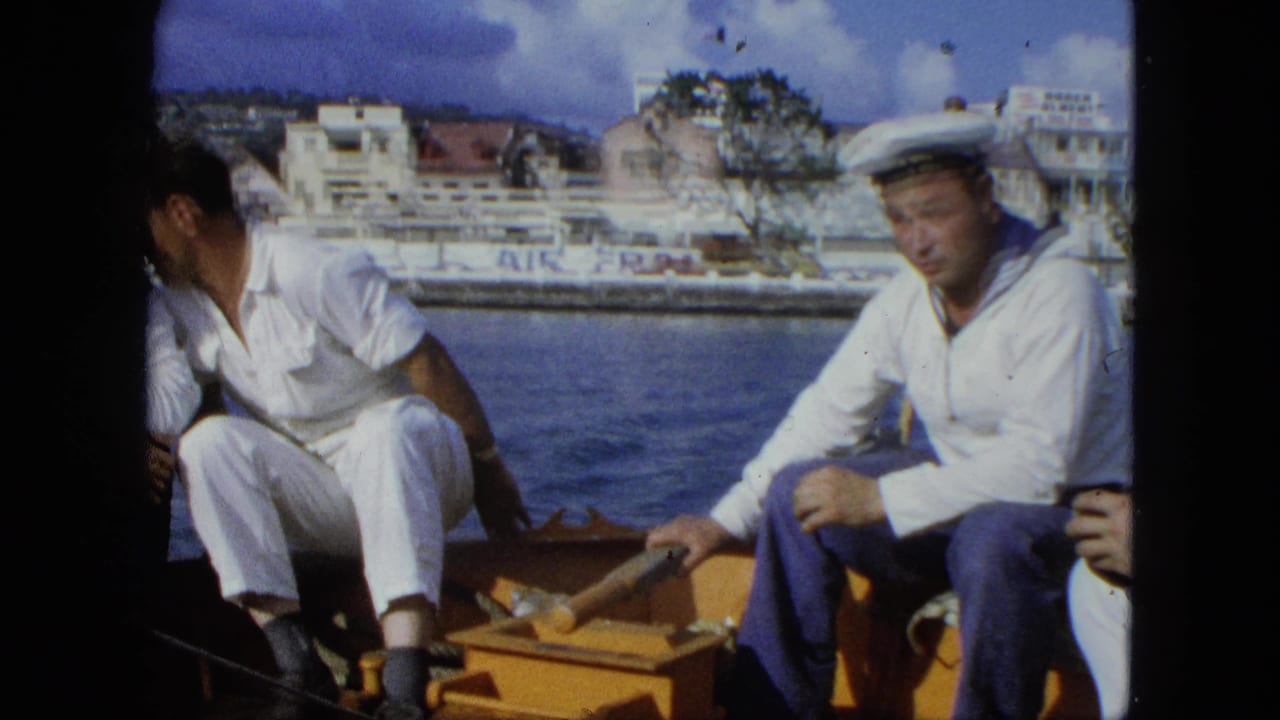 Men in white uniforms sit on a small boat with shoreline buildings behind them.