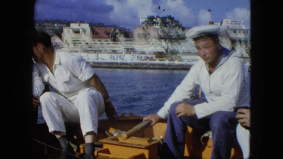 Men in sailor-style clothing sit aboard a small boat in the United States Virgin Islands, with waterfront buildings and Air France lettering behind them.
