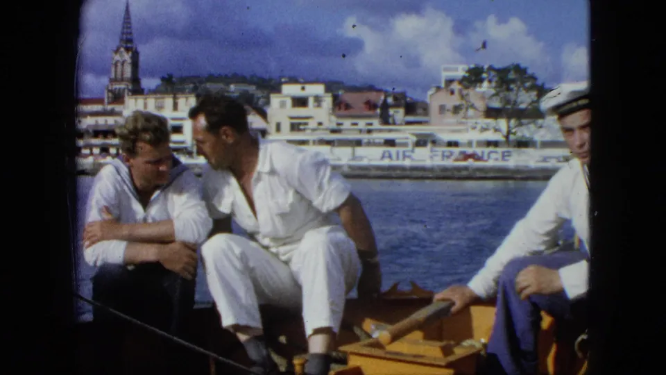 Three men, two in white sailor-style uniforms, sit on a small boat with harbor buildings, a spire, and Air France lettering in the background.