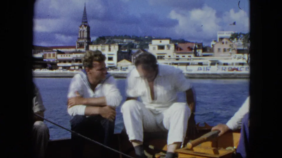 Two men sit and lean toward each other on a small boat, with United States Virgin Islands waterfront buildings and Air France lettering visible behind them.