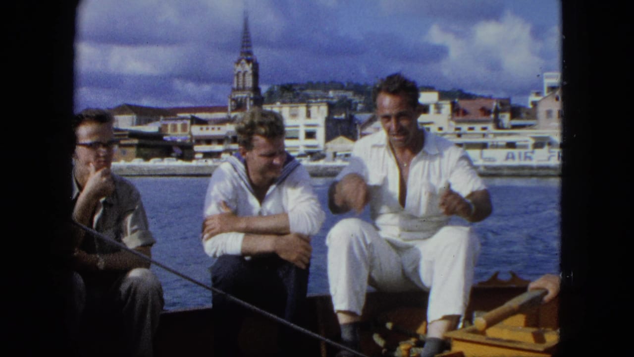 Three adult men sit on a boat in 1964 U.S. Virgin Islands footage, with harbor water and a steepled waterfront town behind them.