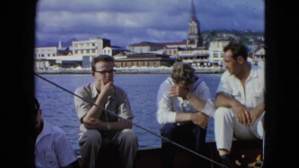 Several adult men are partly visible on a boat in 1964 U.S. Virgin Islands footage, with a harbor town and steeple across the water.