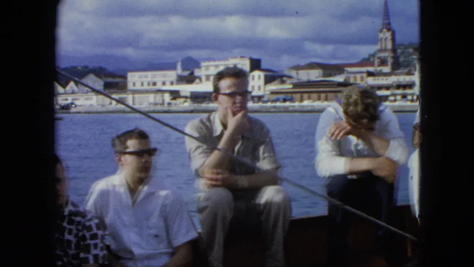 Adult men sit on a boat in 1964 U.S. Virgin Islands home-movie footage, with blue harbor water and low town buildings behind them.