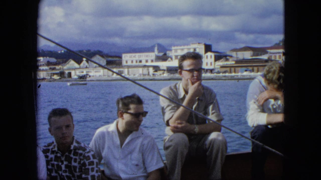 Young men sitting along a boat with water and a coastal town behind them.