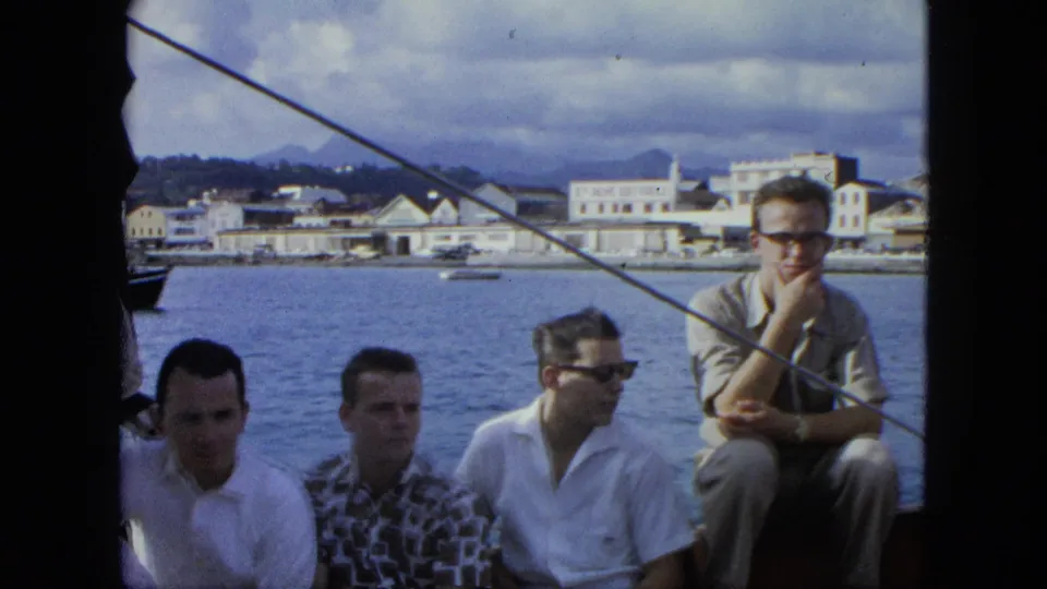 Men in casual shirts sit close together on a boat in the U.S. Virgin Islands, with harbor buildings and hills visible beyond the water.