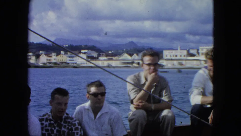 1960s 8mm frame showing men seated on a boat near a coastal town, partly obscured by film edge and boat rigging.