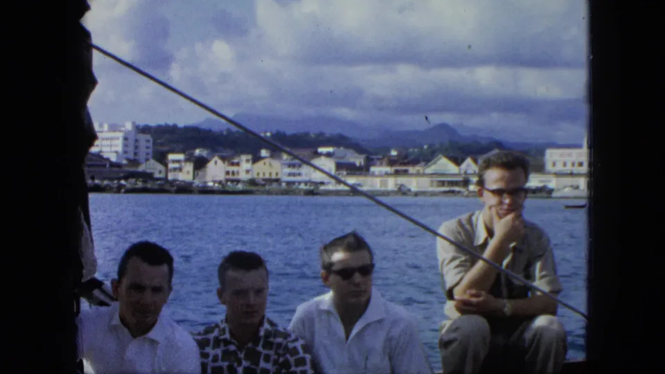 Final frame of young men sitting along a boat in a 1964 U.S. Virgin Islands harbor scene, with waterfront buildings under a cloudy sky.