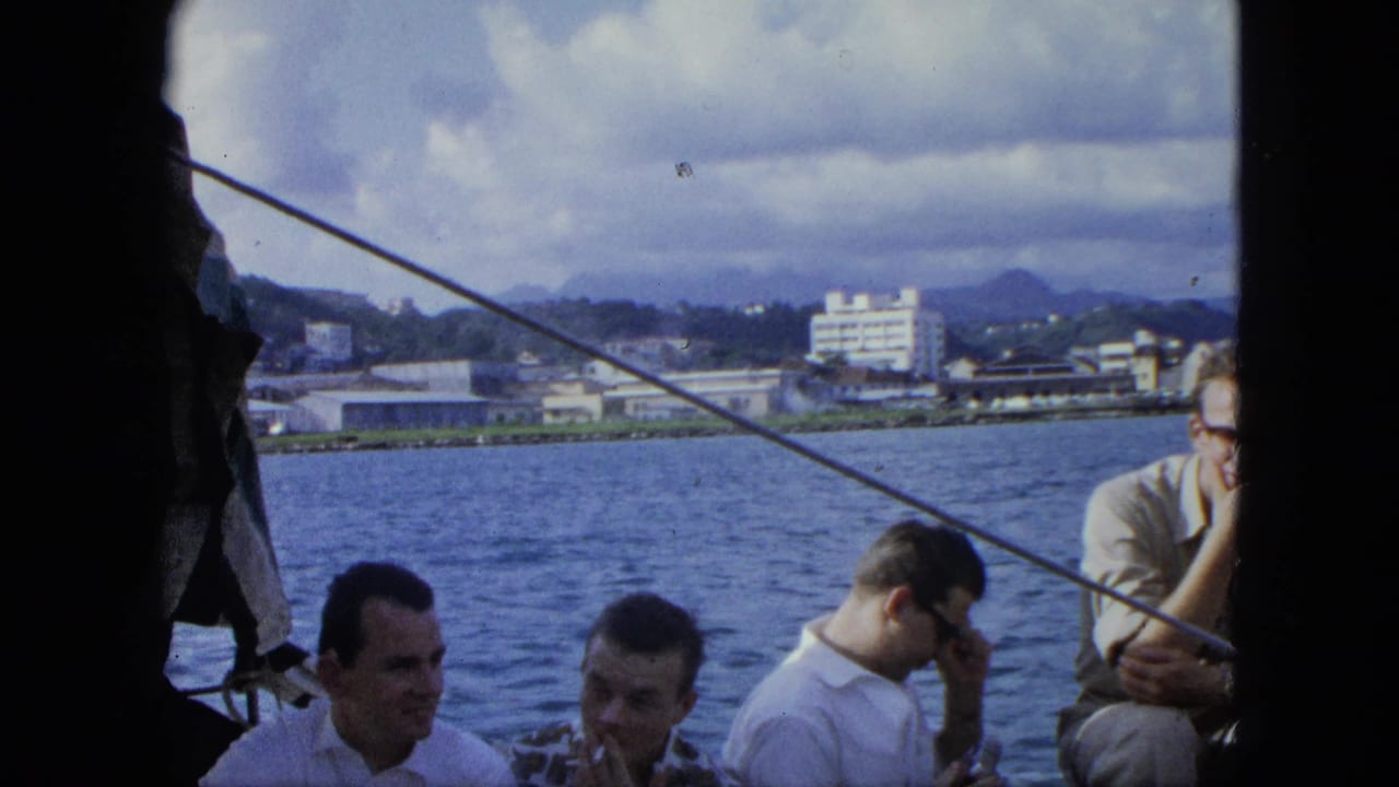 Men seated on a boat with water, shoreline buildings, hills, and cloudy sky behind them.
