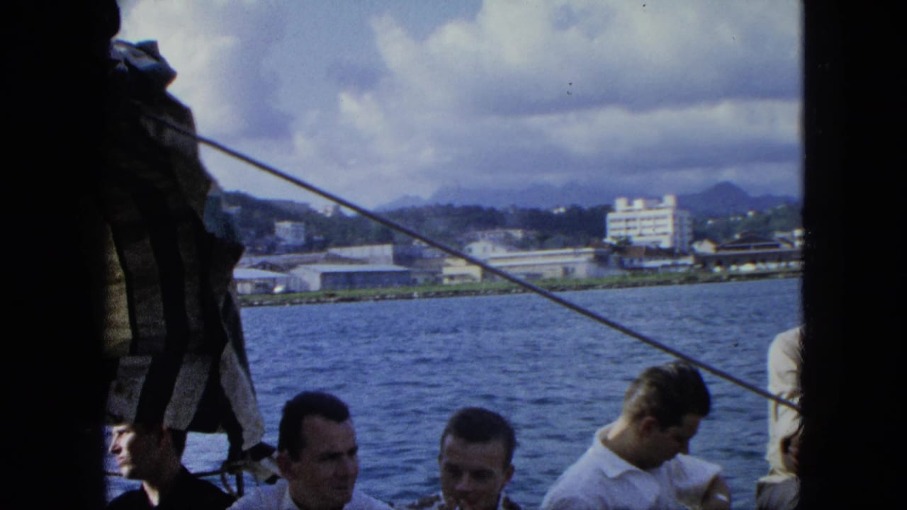 Men in casual 1960s shirts sit on a boat in U.S. Virgin Islands travel footage, with water and shoreline buildings behind them.