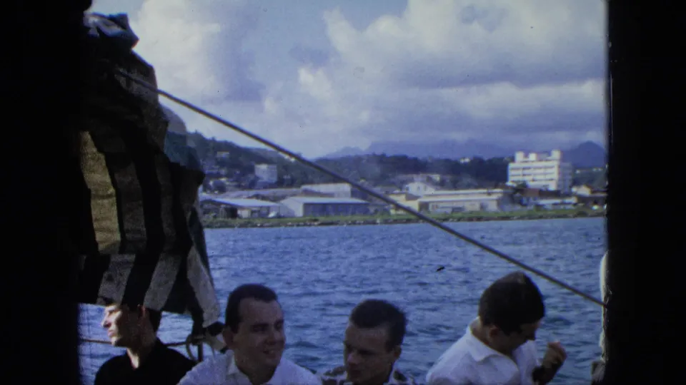 A handheld 8mm frame shows men aboard a boat, partly obscured by canvas or sail material, with U.S. Virgin Islands shoreline in back.