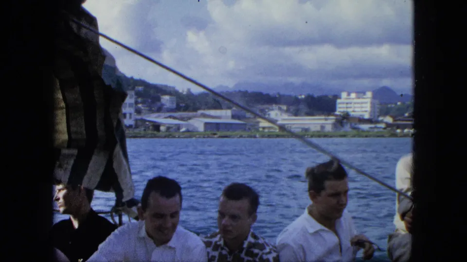 Several adult men on a boat appear in 1964 U.S. Virgin Islands home-movie footage, with water, waterfront buildings, and hills behind them.