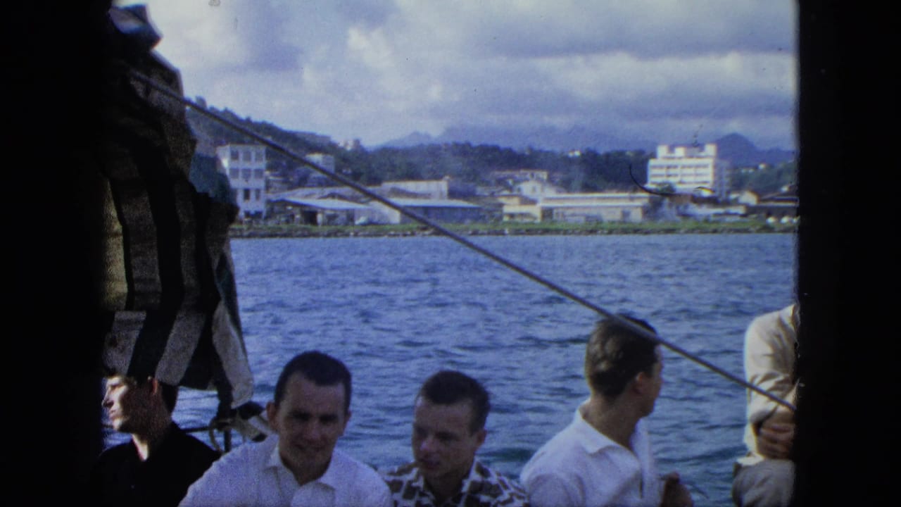 1964 USA Virgin Islands home-movie frame showing men aboard a boat as a coastal town and hills pass in the background.