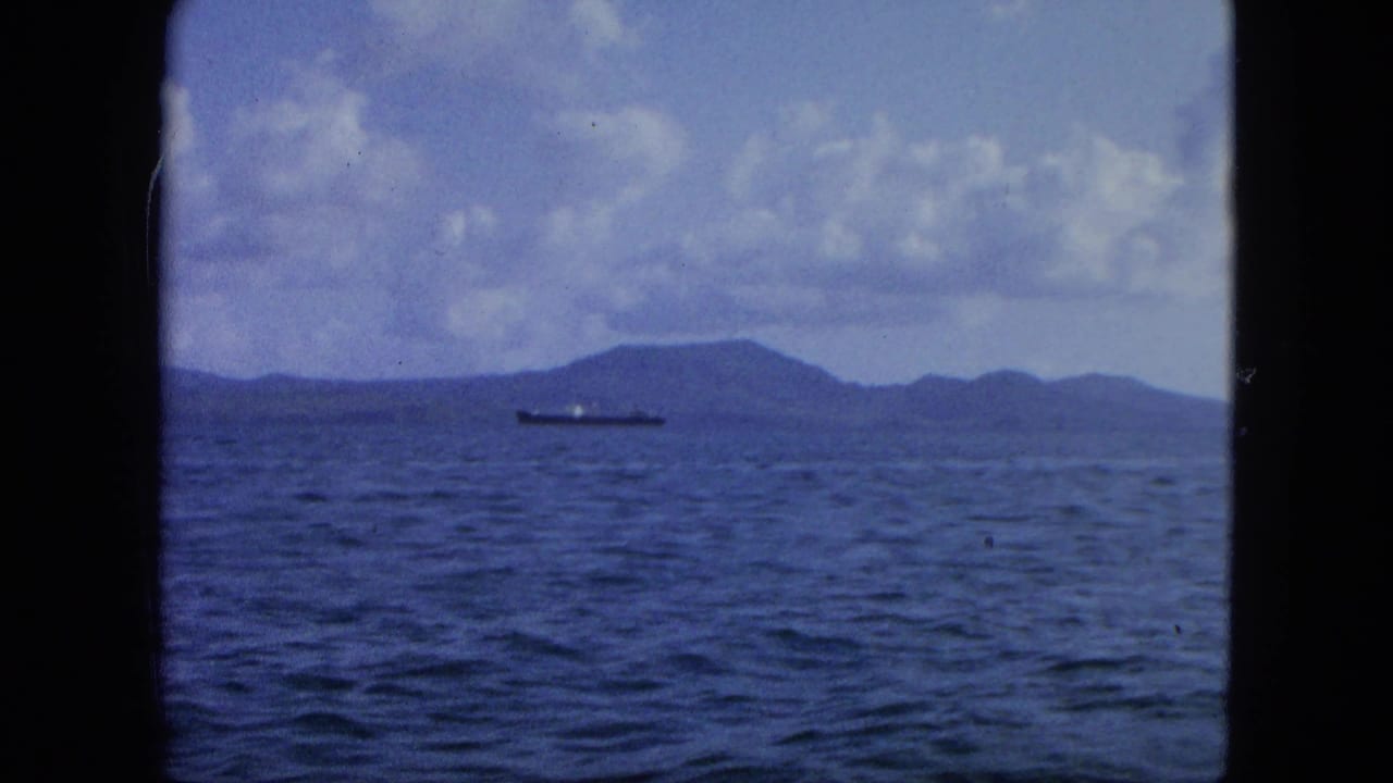 Open water with a distant cargo ship and low islands under a cloudy sky.