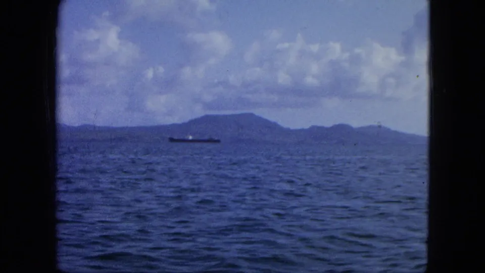 Vintage 8mm view of open water, a distant cargo ship, and low island hills during a 1964 USA Virgin Islands boat trip.