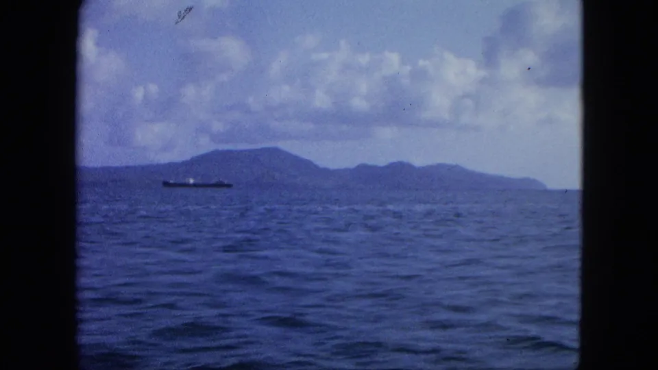 1964 USA Virgin Islands 8mm frame with a cargo ship offshore, low island hills, and dark film-gate borders.