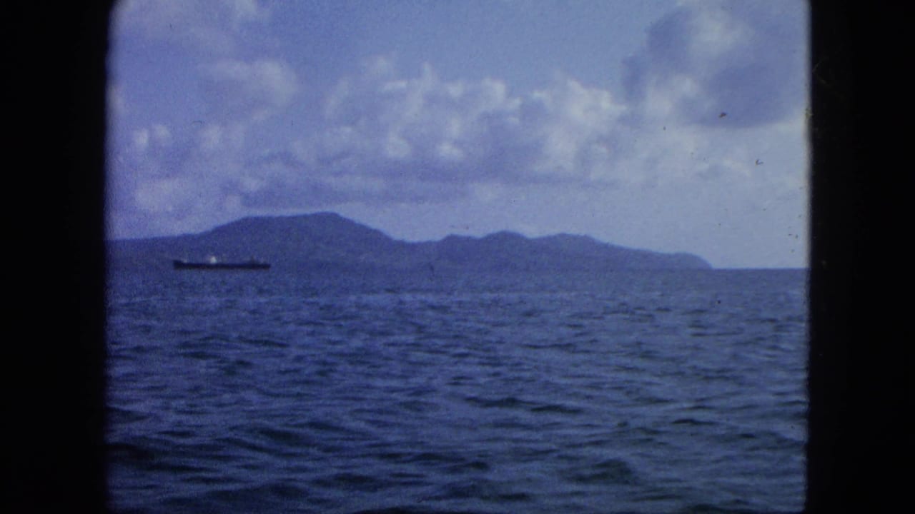 Ocean horizon with distant island, cloudy sky, and a cargo ship near the left side.