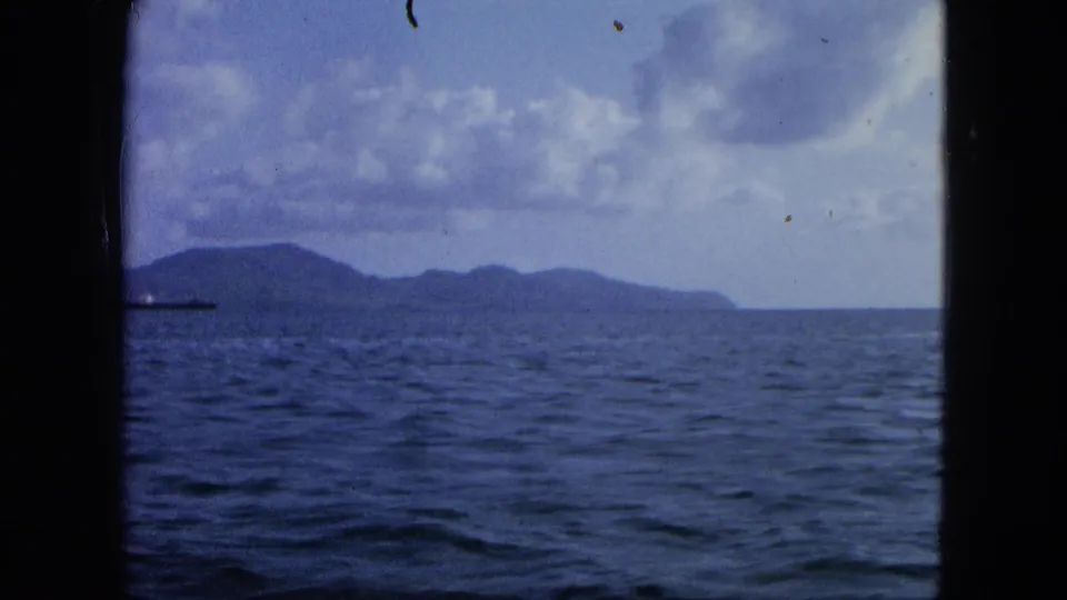 1964 8mm maritime view of open water, island shoreline, clouds, and a cargo ship near the frame edge.
