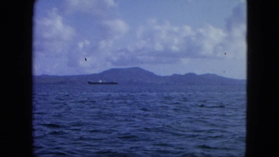 Vintage 8mm view from a vessel showing rippled sea, distant island, and cargo ship under a cloudy sky.