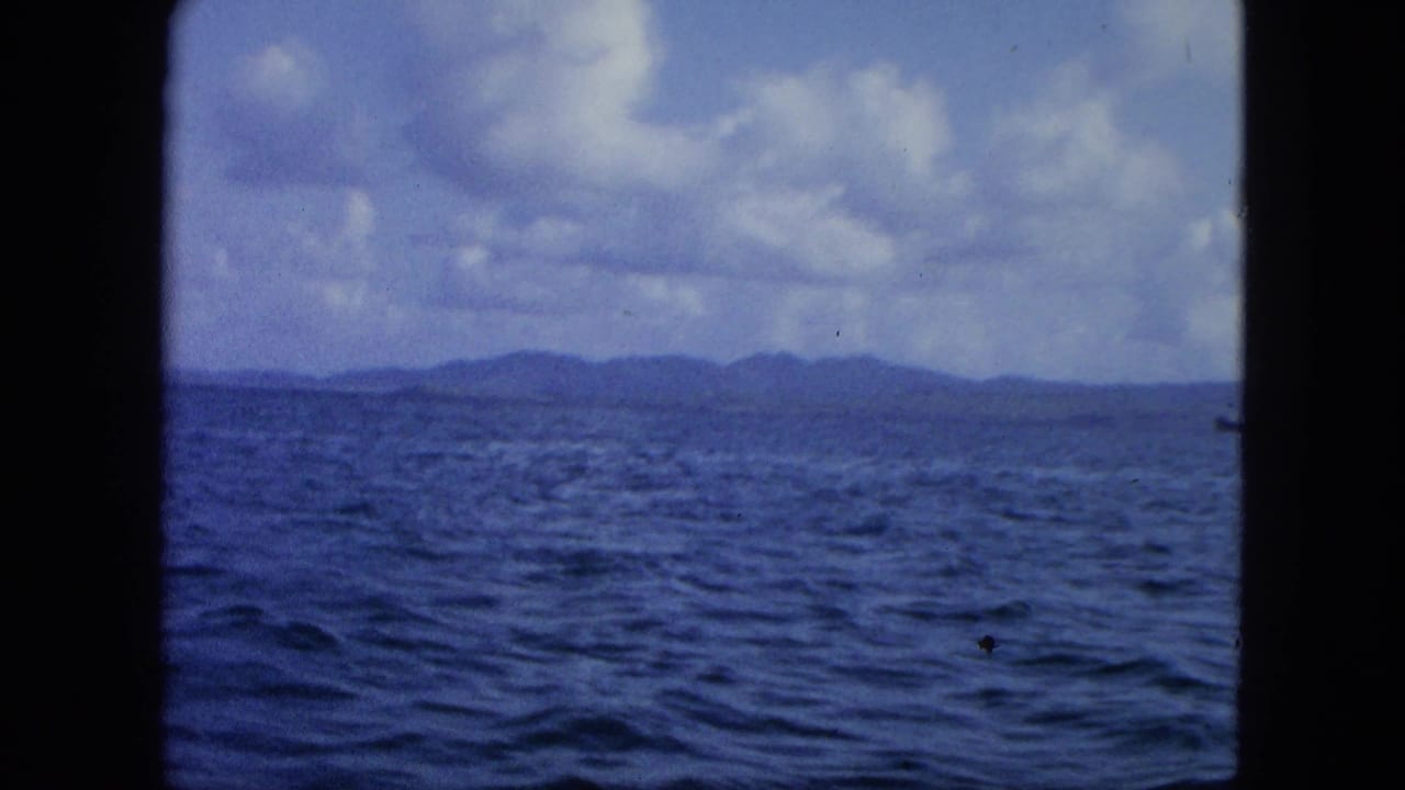 Open ocean and low island horizon beneath a cloudy blue sky, framed by dark film borders.