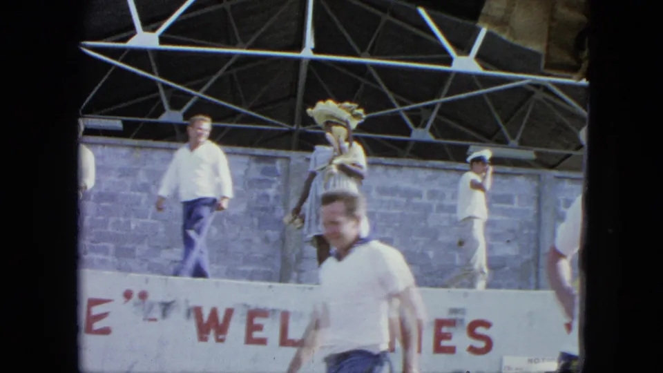 1964 Venezuela home-movie frame showing adults on a stage, including a woman in a wide-brimmed hat, near partial WELCOME lettering.