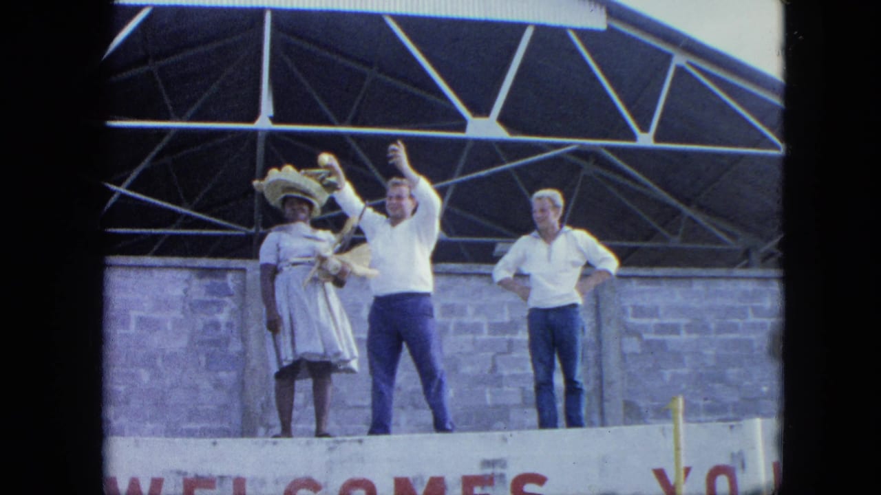 Three adults on a stage, with two people waving near a partial WELCOME sign.