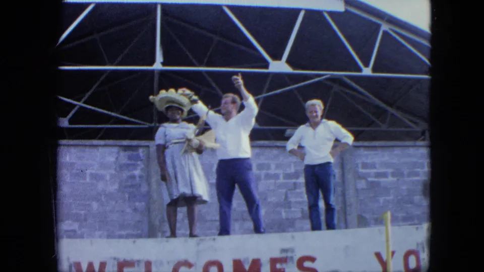 1964 Venezuela home-movie still showing three adults on a stage, with a woman in a white dress and a man raising his hand.
