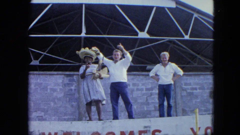 1964 Venezuela frame with three adults on a raised stage, partial WELCOME lettering below, and a man waving beside a woman in a hat.