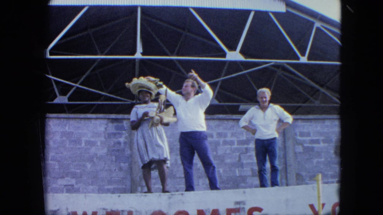 Adults stand on a stage, including a woman in a wide-brimmed hat and two men in white shirts.