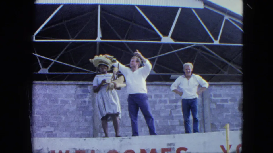 1964 home-movie frame showing adults on a raised stage, with a woman in a white dress and hat beside two men under roof trusses.