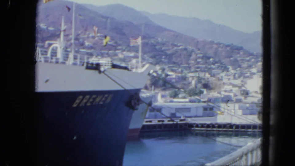 1964 travel footage frame showing the Bremen at port, with harbor water, dock structures, hillside buildings, and mountains in the background.