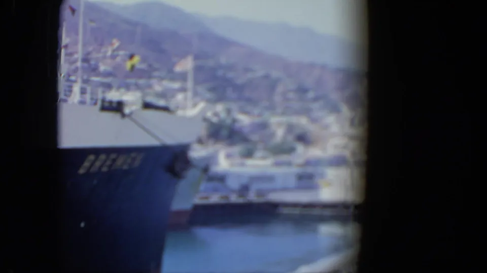 Soft 8mm frame of the Bremen docked at a harbor, with water, pier structures, a hillside town, and mountains visible beyond.