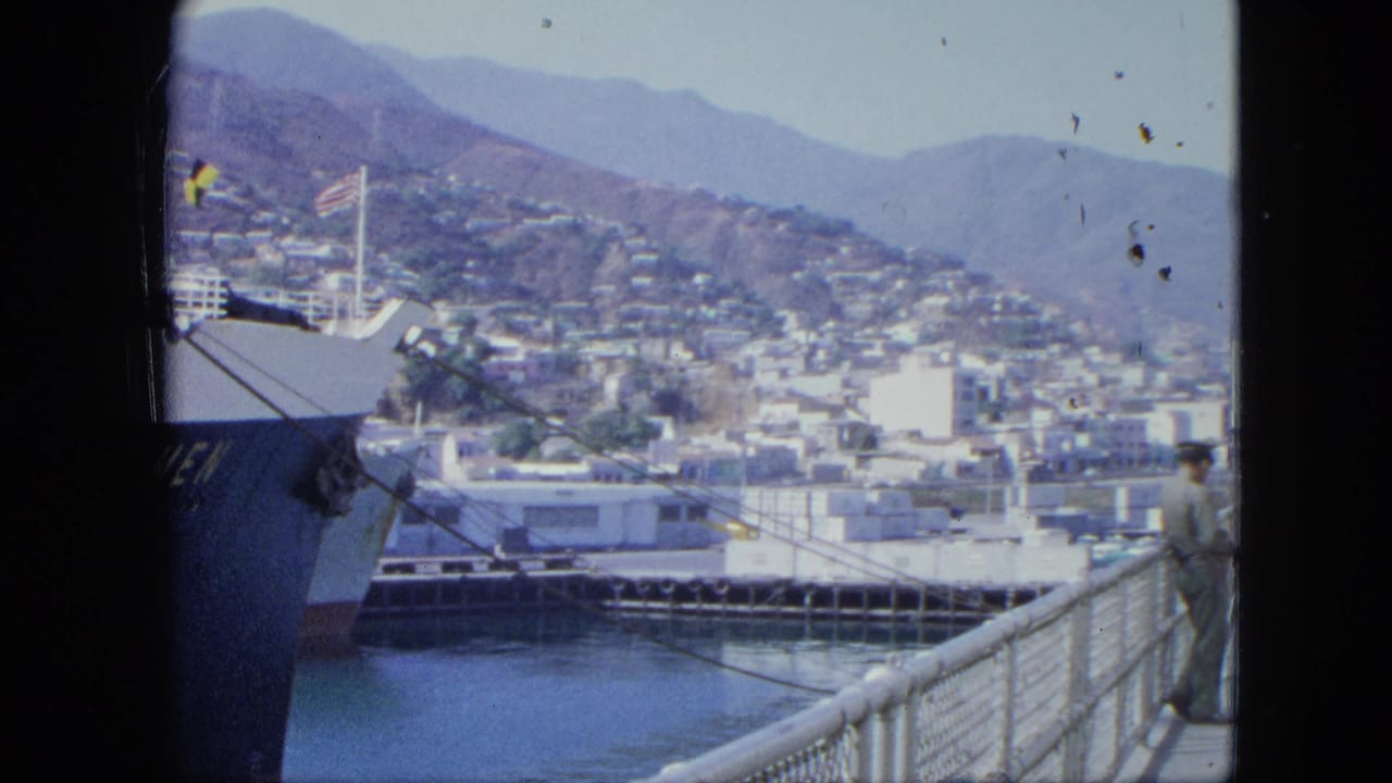 1964 8mm dockside view in Venezuela with a moored ship, flag, harbor railings, hillside city, mountains, and a uniformed man at right.