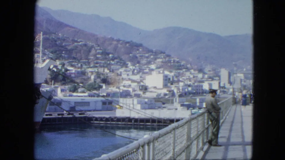 1964 Venezuela harbor frame showing a ship at left, metal dock railing, a uniformed man on the pier, hillside buildings, and mountains beyond.