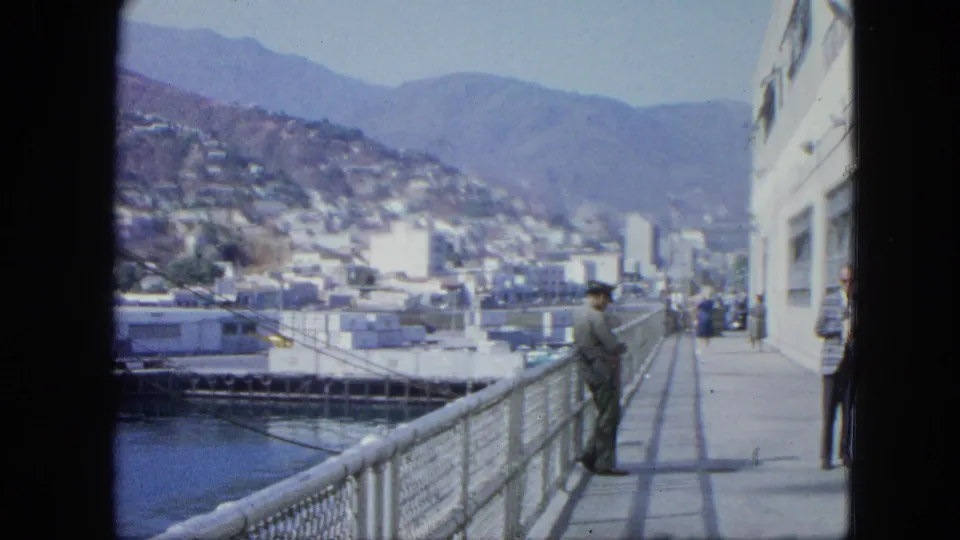 1964 dockside frame in Venezuela with a white ship wall, uniformed man by the railing, harbor basin, hillside city, and mountains in the distance.