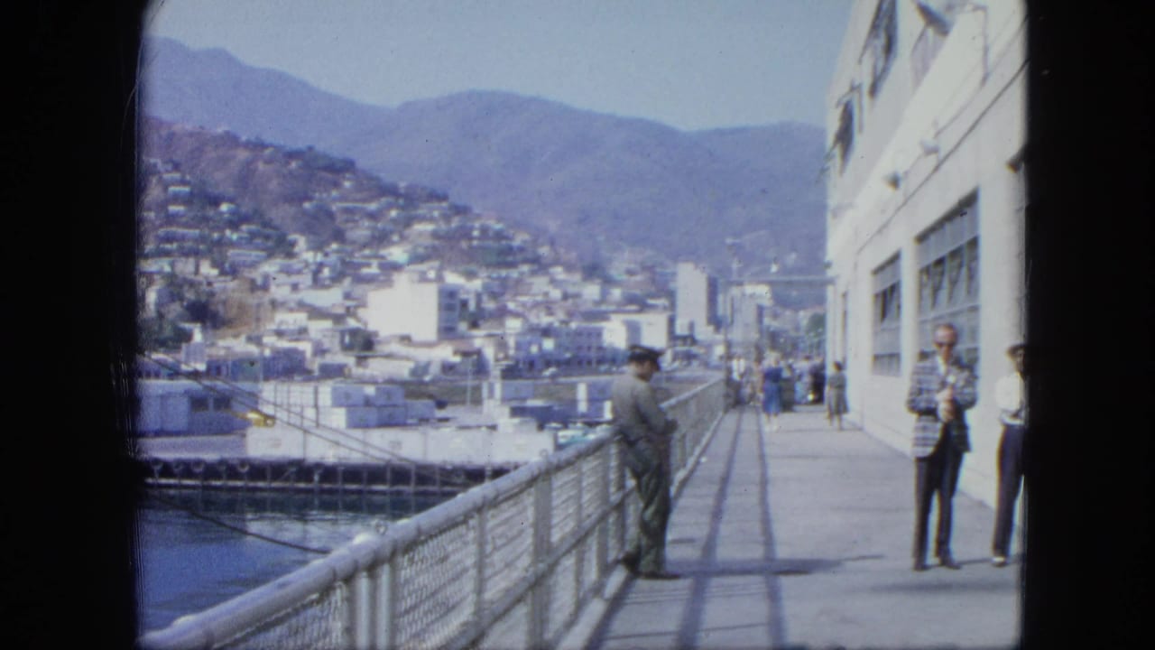 1964 Venezuela harbor view with adults on a pier, a white waterfront building, water, and a hillside city in the background.