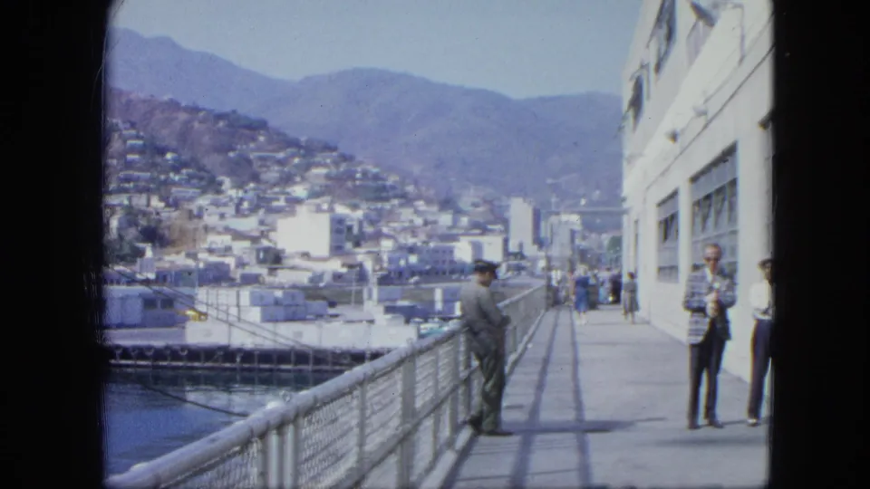1964 Venezuela harbor view with adults on a pier, a white waterfront building, water, and a hillside city in the background.