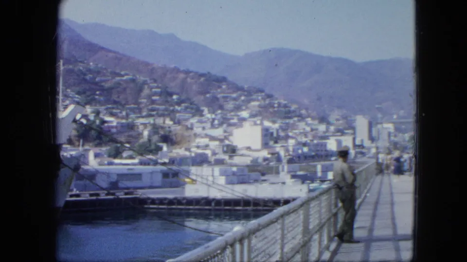 The camera frames the pier railing, a man in period clothing, moored harbor craft, and the dense mountain-backed city in Venezuela.